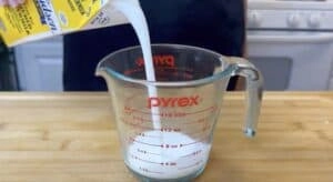 Pouring buttermilk into a glass measuring cup on a wooden countertop in a kitchen setting.